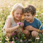 Insekten von A bis Z Zwei kleine Kinder hocken auf einer blumenreichen Wiese und beobachten lächelnd einen Schmetterling, der im Sonnenlicht auf einer rosa Blüte sitzt – ein perfekter Moment des Staunens aus der Welt von „Insekten von A bis Z“.