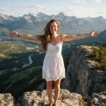 Zitate über Freiheit & Unabhängigkeit Eine Frau in einem weißen Sommerkleid steht mit ausgestreckten Armen auf einer felsigen Klippe und lächelt, während sie unter klarem Himmel auf eine malerische Berglandschaft mit einem Fluss und einem Wald im Tal blickt – ein Sinnbild für den Geist der Zitate über Freiheit und Unabhängigkeit.