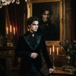Zitate von Oscar Wilde Ein Mann in einem dunklen Anzug im viktorianischen Stil steht in einem prunkvollen Raum und hält ein Buch mit Zitaten von Oscar Wilde in der Hand; hinter ihm hängt das Porträt einer ähnlich gekleideten Gestalt mit einem finsteren Lächeln, umgeben von Kerzen und antikem Dekor.