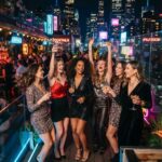 Zitate über Wochenende & Feierabend Sechs Frauen in eleganten Abendkleidern lachen und feiern bei einem Drink in einer lebhaften Dachterrassenbar am Abend und zitieren dabei ihre Lieblingszitate zum Thema „Wochenende & Feierabend“, während im Hintergrund die hellen Lichter der Stadt und die Leuchtreklamen leuchten.