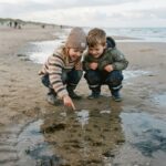 Krebstiere von A bis Z Zwei kleine Kinder hocken an einem Sandstrand, blicken nach unten und lächeln, während eines von ihnen auf etwas in einem flachen Gezeitentümpel zeigt – vielleicht entdecken sie gerade faszinierende Krebstiere von A bis Z. Der Himmel ist bewölkt, und im Hintergrund ist das Meer zu sehen.