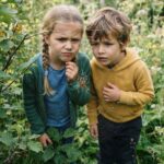 Spinnentiere von A bis Z Zwei kleine Kinder betrachten aufmerksam eine große Spinne, die inmitten grüner Pflanzen im Freien in der Mitte ihres Netzes sitzt. Während das Mädchen skeptisch wirkt und der Junge neugierig, sind beide fasziniert von ihrer Begegnung in der freien Natur mit einem der „Spinnentiere von A bis Z“.